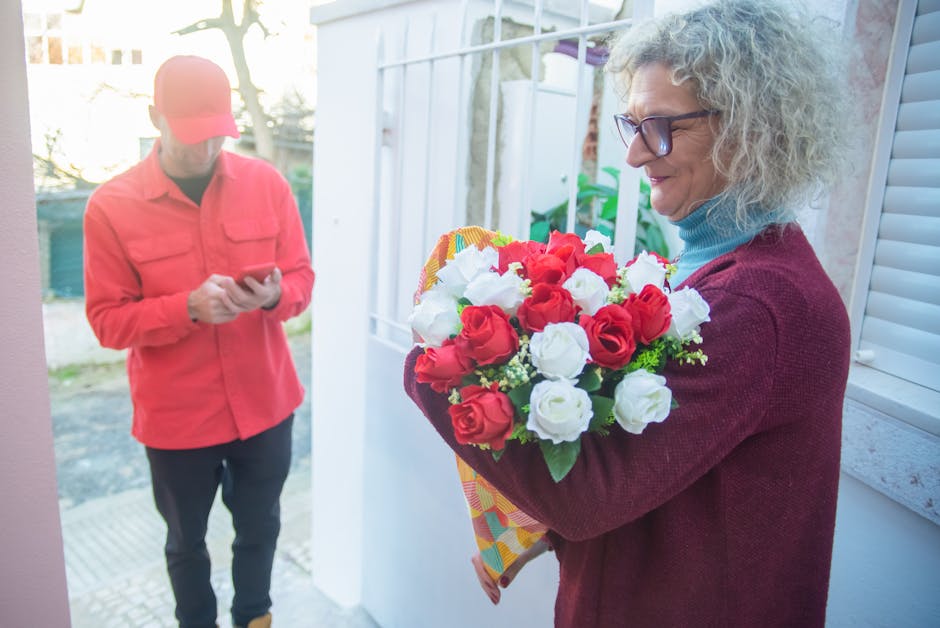 A woman with curly gray hair and glasses holds a vibrant floral bouquet featuring red and white roses, accented with small green foliage, wrapped in colorful patterned paper. She stands near a glass d