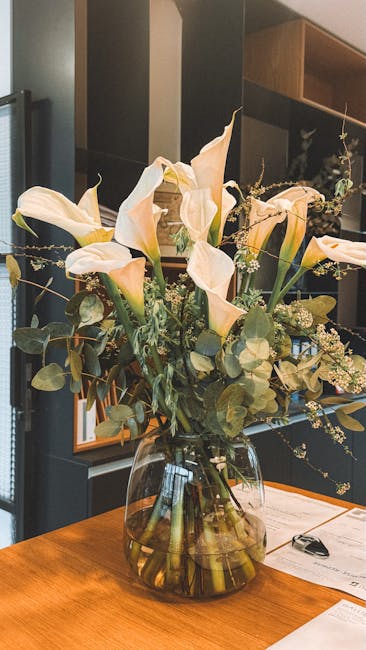 A floral arrangement featuring white calla lilies with elegant, trumpet-shaped petals and long green stems, complemented by sprigs of small white filler flowers and eucalyptus leaves with rounded silv