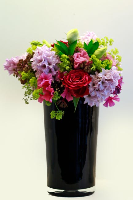 A vibrant floral bouquet arranged in a tall, glossy black vase, featuring a mixture of fresh flowers including a prominent red rose at the center, surrounded by soft pink and lavender stock, white tul