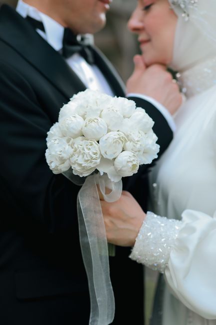 Close-up of a bride and groom in wedding attire, with the bride holding a bouquet of white peonies and roses wrapped with white ribbon. The groom is wearing a classic black tuxedo with a bow tie, and