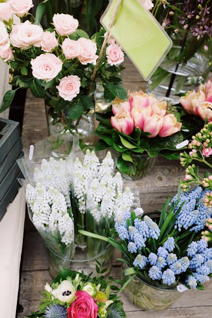 A display of fresh flowers arranged in clear glass vases on a wooden surface, featuring light pink spray roses with dark green foliage, pink and cream tulips with large, smooth petals, clusters of whi