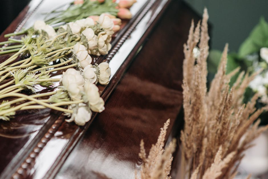 A close-up of a floral arrangement placed on a polished wooden surface, featuring a freshly prepared bouquet of predominantly white flowers with soft petals and green stems, complemented by dried beig