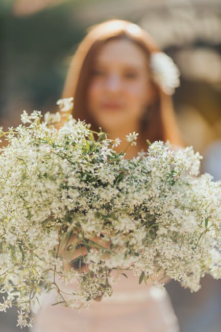 A woman with red hair, wearing a white dress and a white floral hair accessory, holds a large bouquet of white flowers with small, delicate petals and green stems in front of her. The bouquet is arran
