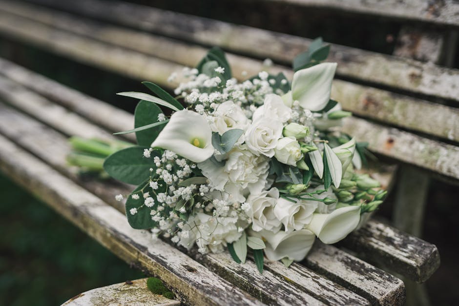 A bouquet of fresh white flowers, including calla lilies, roses, and lisianthus, accented with eucalyptus and baby's breath, arranged in a loose, elegant style. The bouquet is wrapped with white flora