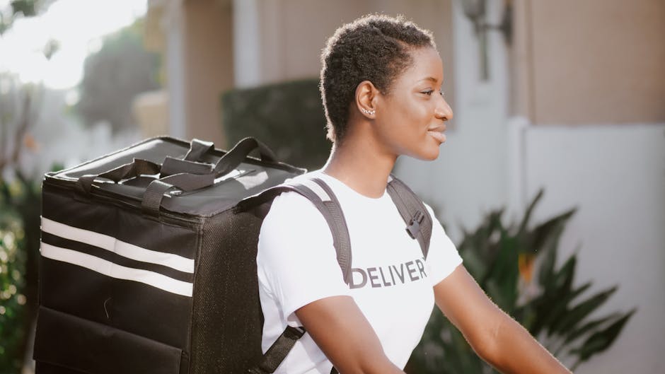 A young woman with short, curly hair and medium skin tone is captured in profile, wearing a white T-shirt with the word 'DELIVERY' printed on it. She is outdoors in natural light, standing next to a m