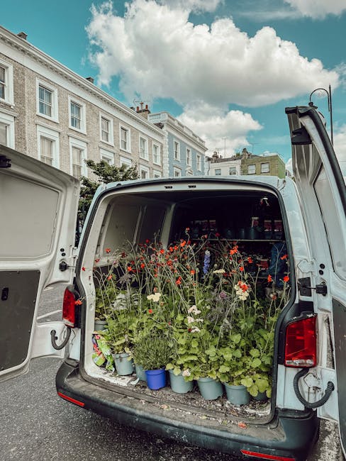 The back of a white delivery van open to reveal an array of freshly picked potted flowers and blooming plants, including tall red and white poppies with delicate petals and green stems, arranged on sh